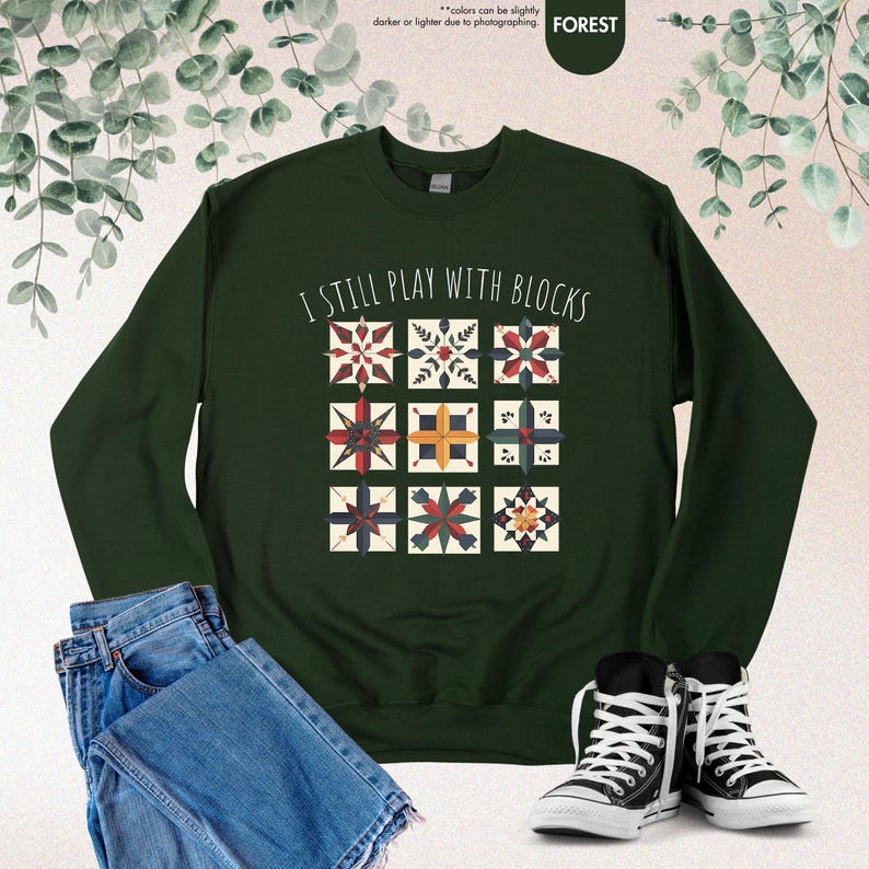 May include: A dark green sweatshirt with the text "I still play with blocks" in white. The sweatshirt features a design of 12 colorful patchwork blocks.