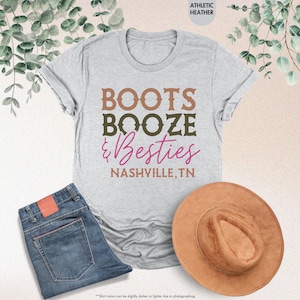 Pu&ograve; includere: Una maglietta grigio chiaro con le parole "BOOTS BOOZE & Besties NASHVILLE, TN" in marrone, verde oliva e rosa. La maglietta &egrave; abbinata a jeans blu e un cappello marrone.