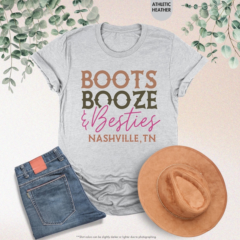 Pu&ograve; includere: Una maglietta grigio chiaro con le parole "BOOTS BOOZE & Besties NASHVILLE, TN" in marrone, verde oliva e rosa. La maglietta &egrave; abbinata a jeans blu e un cappello marrone.