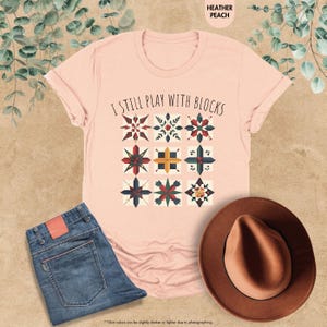 May include: A pink t-shirt with the text "I still play with blocks" and a design of colorful quilt blocks.