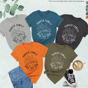 May include: Five short-sleeved t-shirts in heather grey, teal, dark grey, olive, and orange. Each shirt features a white graphic with the text "PARKER FAMILY ROAD TRIP 2026". The shirts are displayed with a pair of jeans, a cup of coffee, and a pair of black trainers.