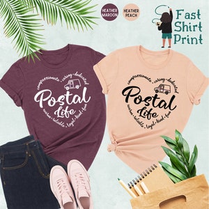 May include: Two t-shirts, one heather maroon and one heather peach, with a white graphic design that says "Postal Life" in a circle with a mail truck and the words "compassionate, caring, dedicated, warm, reliable, loyal, kind, fun".