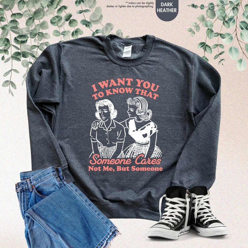 May include: Dark heather gray sweatshirt with the text "I WANT YOU TO KNOW THAT Someone Cares Not Me, But Someone" and a graphic of two women. The sweatshirt is paired with blue jeans and black and white sneakers.