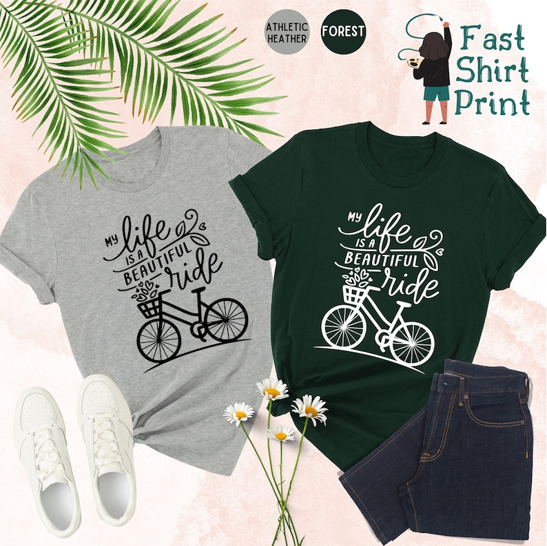 May include: Two t-shirts with the text "My life is a beautiful ride" and a bicycle graphic. The shirt on the left is heather grey with black text and graphic. The shirt on the right is forest green with white text and graphic.