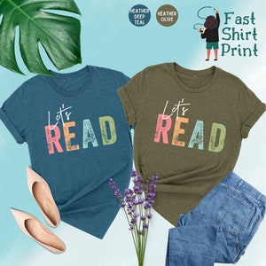May include: Two t-shirts with the text "Let's Read" in colorful letters. The shirt on the left is teal blue and the shirt on the right is olive green.