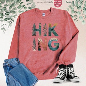 Pode incluir: Uma camisa de moletom vermelho escarlate com o termo "HIKING" em letras grandes. As letras s&atilde;o compostas por ilustra&ccedil;&otilde;es de &aacute;rvores, montanhas e outras cenas da natureza.