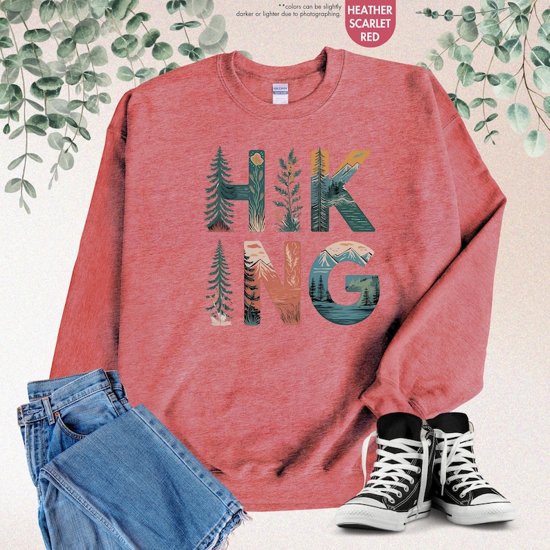 Pode incluir: Uma camisa de moletom vermelho escarlate com o termo "HIKING" em letras grandes. As letras s&atilde;o compostas por ilustra&ccedil;&otilde;es de &aacute;rvores, montanhas e outras cenas da natureza.