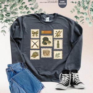 K&ouml;nnte beinhalten: Dunkelgraues Sweatshirt mit Jagd-Postwertzeichen-Designs. Die Briefmarken zeigen eine Schleife, eine Schrotflinte, einen Jagdhund mit einer Ente, eine Ente, einen Jagdhut, eine Jagdbox und einen bambusartigen Gegenstand. Das Sweatshirt wird mit blauen Jeans und schwarz-wei&szlig;en Turnschuhen kombiniert.