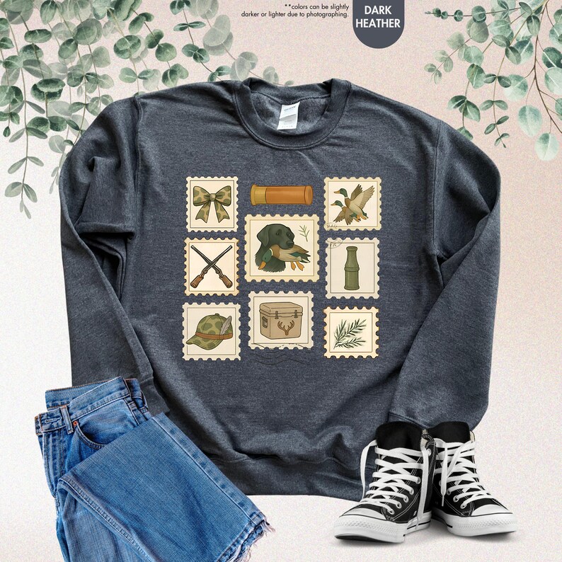 K&ouml;nnte beinhalten: Dunkelgraues Sweatshirt mit Jagd-Postwertzeichen-Designs. Die Briefmarken zeigen eine Schleife, eine Schrotflinte, einen Jagdhund mit einer Ente, eine Ente, einen Jagdhut, eine Jagdbox und einen bambusartigen Gegenstand. Das Sweatshirt wird mit blauen Jeans und schwarz-wei&szlig;en Turnschuhen kombiniert.