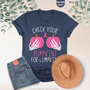以下が含まれることがあります： 「CHECK YOUR PUMPKINS FOR LUMPKINS」の文字が入ったネイビーブルーのTシャツ。ピンクのリボンと、骸骨の手が付いた2つのピンクのかぼちゃがデザインされています。青いジーンズと茶色の帽子も画像に写っています。