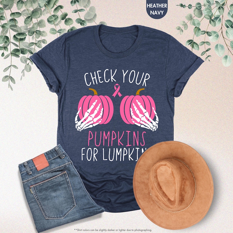 以下が含まれることがあります： 「CHECK YOUR PUMPKINS FOR LUMPKINS」の文字が入ったネイビーブルーのTシャツ。ピンクのリボンと、骸骨の手が付いた2つのピンクのかぼちゃがデザインされています。青いジーンズと茶色の帽子も画像に写っています。