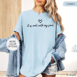 K&ouml;nnte beinhalten: Hellblaues T-Shirt mit dem handgeschriebenen Schriftzug "it is well with my soul" und einem kleinen Herz dar&uuml;ber. Das Shirt hat hochgekrempelte &Auml;rmel und ist aus weichem Stoff. Eine Jeansjacke ist ebenfalls im Bild zu sehen.