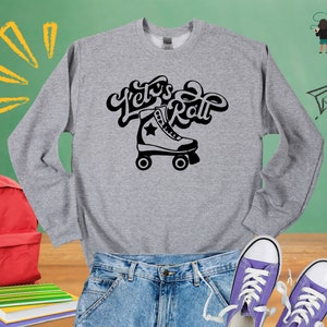 May include: A grey sweatshirt with the text "Let's Roll" in black, featuring a black and white roller skate graphic. The sweatshirt is displayed on a wooden surface with a red backpack, a pair of blue jeans, and two purple sneakers.