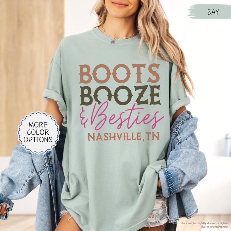 Pu&ograve; includere: T-shirt verde chiaro con le parole "BOOTS BOOZE & Besties" in marrone, verde oliva e rosa. Il testo "NASHVILLE, TN" &egrave; in basso. La maglietta ha le maniche arrotolate ed &egrave; indossata con una giacca di jeans.