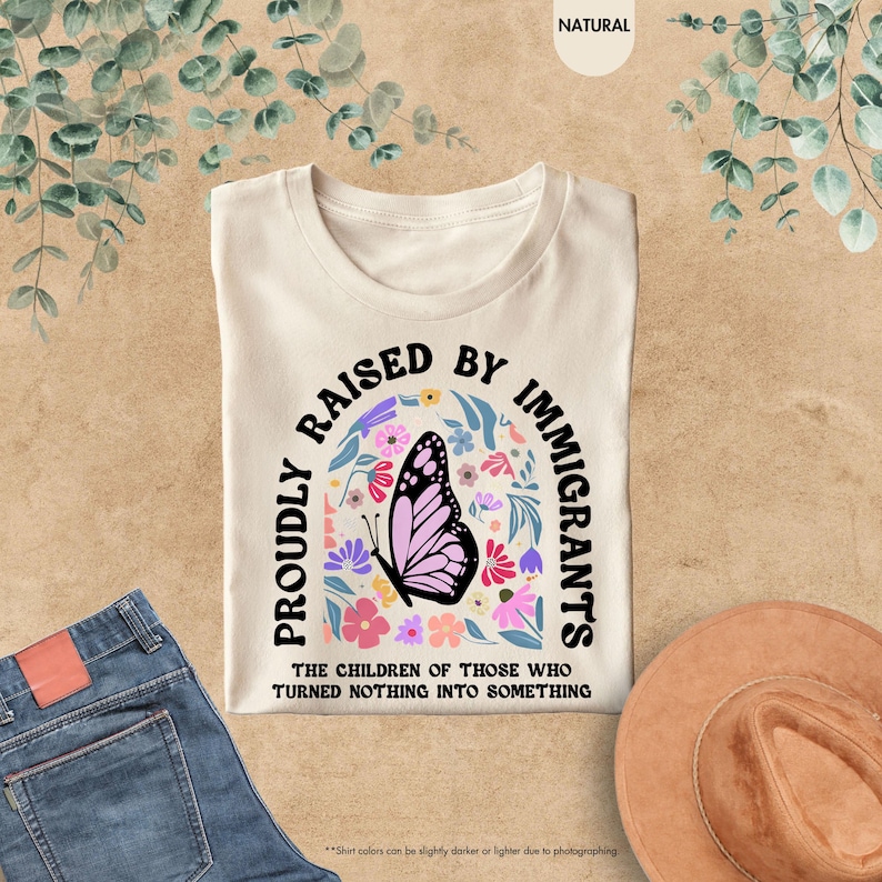 May include: Cream-colored t-shirt with the text "PROUDLY RAISED BY IMMIGRANTS" in black, arched above a butterfly and floral design. The text "THE CHILDREN OF THOSE WHO TURNED NOTHING INTO SOMETHING" is below. The shirt is folded on a neutral surface.