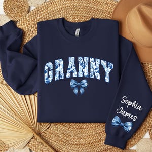 May include: Navy blue sweatshirt with the word "Granny" in white letters with a blue floral pattern. A blue bow is below the word. The left sleeve has the name "Sophia James" in white letters with a blue floral pattern and a blue bow.