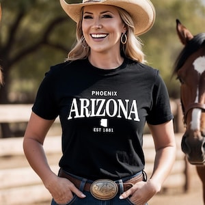 May include: A black t-shirt with the word "ARIZONA" in white block letters, with "PHOENIX" above and "EST 1881" below. The shirt is worn by a person wearing a cowboy hat and a belt with a decorative buckle.