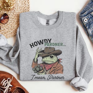 May include: A grey sweatshirt featuring a cartoon frog wearing a cowboy hat and holding a cactus. The text on the sweatshirt reads "Howdy Pardner... I mean, Partner".