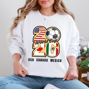 May include: White sweatshirt featuring a graphic for the 2026 World Cup, with the numbers 26 in red and yellow, and a soccer ball. The flags of the USA, Canada, and Mexico are incorporated into the design. The text "USA CANADA MEXICO" is printed below.