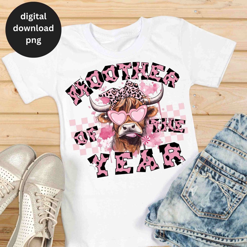 May include: A white t-shirt with a pink and black graphic of a cow wearing heart-shaped sunglasses and a pink bandana. The text "Moother of the Year" is written in pink and black letters.