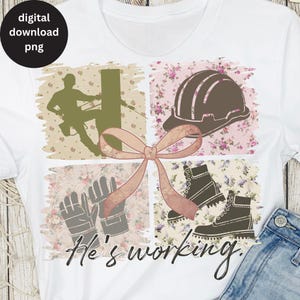 May include: A white t-shirt with a pink bow and a floral design. The design includes a silhouette of a worker on a pole, a hard hat, work gloves, and work boots. The text "He's working" is printed on the shirt.