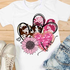 Puede incluir: Una camiseta blanca con un dise&ntilde;o rosa, marr&oacute;n y dorado de tres corazones, un girasol y la palabra "love".