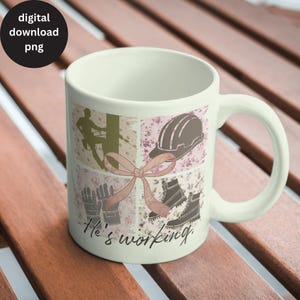May include: White ceramic mug with a collage design featuring a silhouette of a person climbing a pole, a hard hat, work gloves, and boots. The mug has the text "He's working." printed on the bottom.