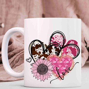 Puede incluir: Taza de cer&aacute;mica blanca con un dise&ntilde;o rosa, marr&oacute;n y dorado de tres corazones, un girasol y la palabra "love".