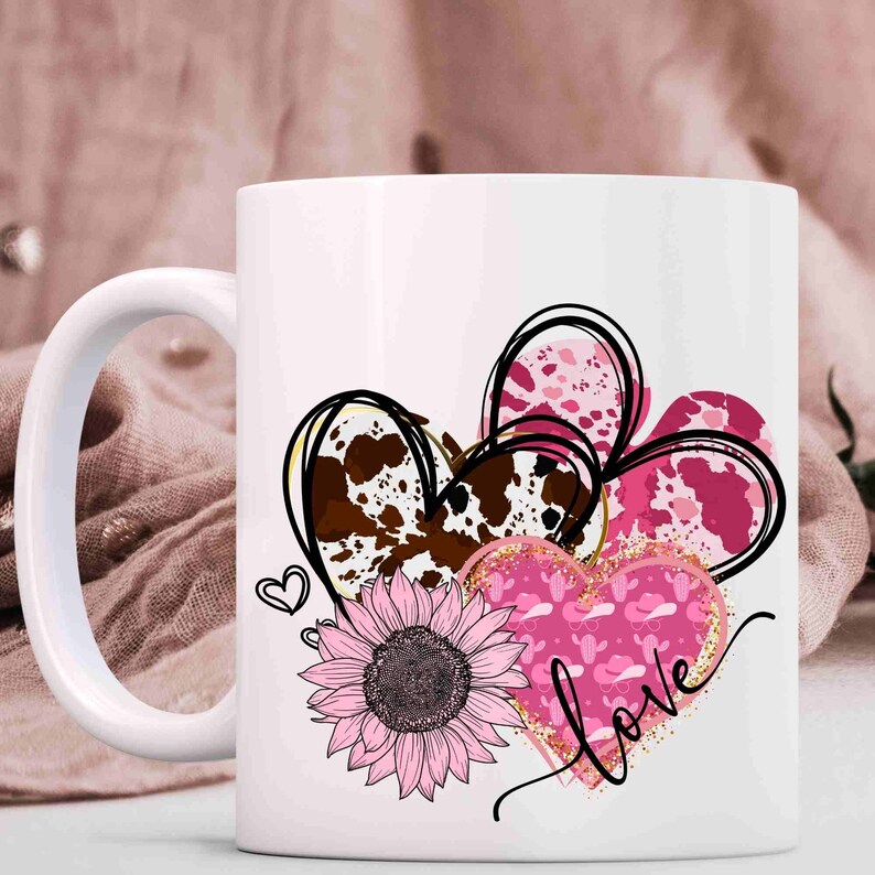Puede incluir: Taza de cer&aacute;mica blanca con un dise&ntilde;o rosa, marr&oacute;n y dorado de tres corazones, un girasol y la palabra "love".