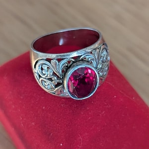 May include: Silver ring with a large, oval, deep red gemstone. The ring has intricate floral patterns around the band. The gemstone is set in a bezel setting. The ring is displayed on a red surface.