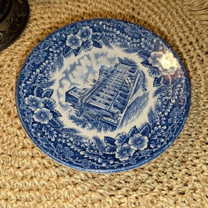 May include: A blue and white decorative plate with a detailed illustration of a building. The plate features a floral border and a central image of a building, likely a landmark. The plate is set on a woven mat.