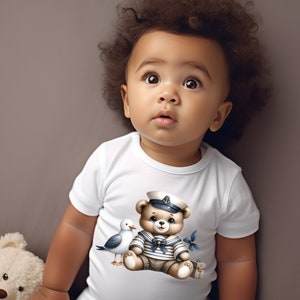 May include: A white baby onesie with a cute cartoon illustration of a brown bear wearing a sailor hat and a blue and white striped shirt. The bear is holding a small suitcase and a seagull is flying next to it.