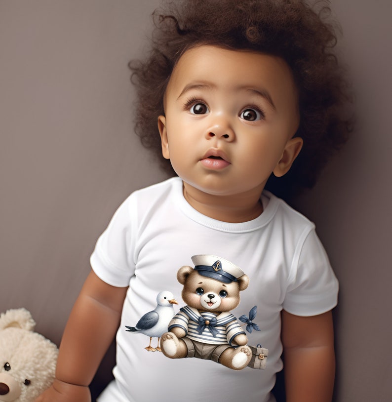 May include: A white baby onesie with a cute cartoon illustration of a brown bear wearing a sailor hat and a blue and white striped shirt. The bear is holding a small suitcase and a seagull is flying next to it.