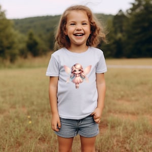 May include: A young girl wearing a light blue t-shirt with a graphic of a fairy with pink wings and a pink dress. The girl is smiling and looking at the camera.