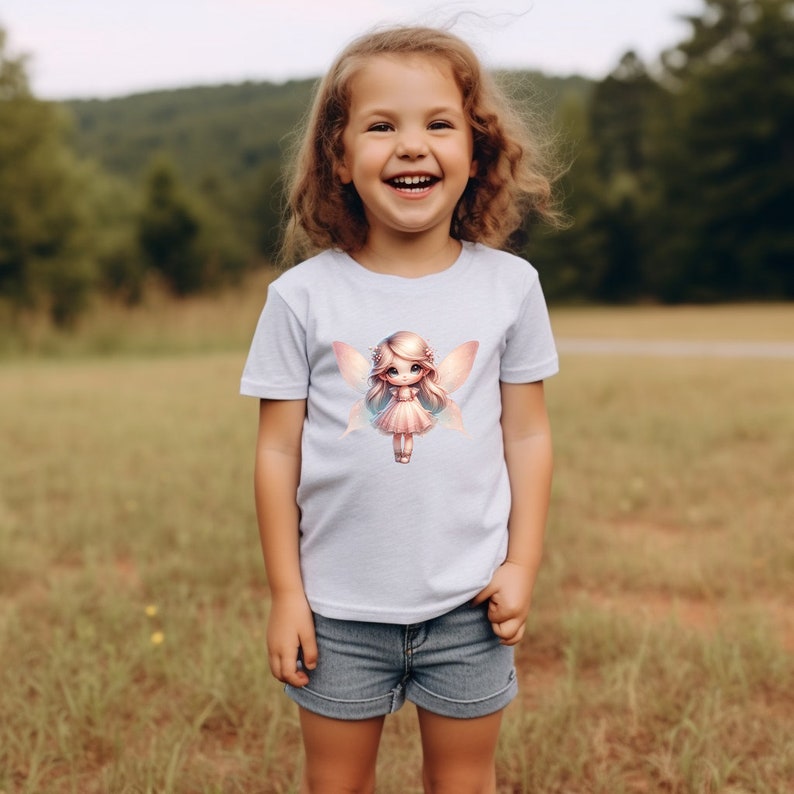May include: A young girl wearing a light blue t-shirt with a graphic of a fairy with pink wings and a pink dress. The girl is smiling and looking at the camera.