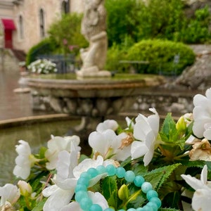 May include: A light blue beaded bracelet with round beads, resting on white flowers and green leaves. The bracelet is in focus, while the background is blurred and features a stone fountain and greenery.
