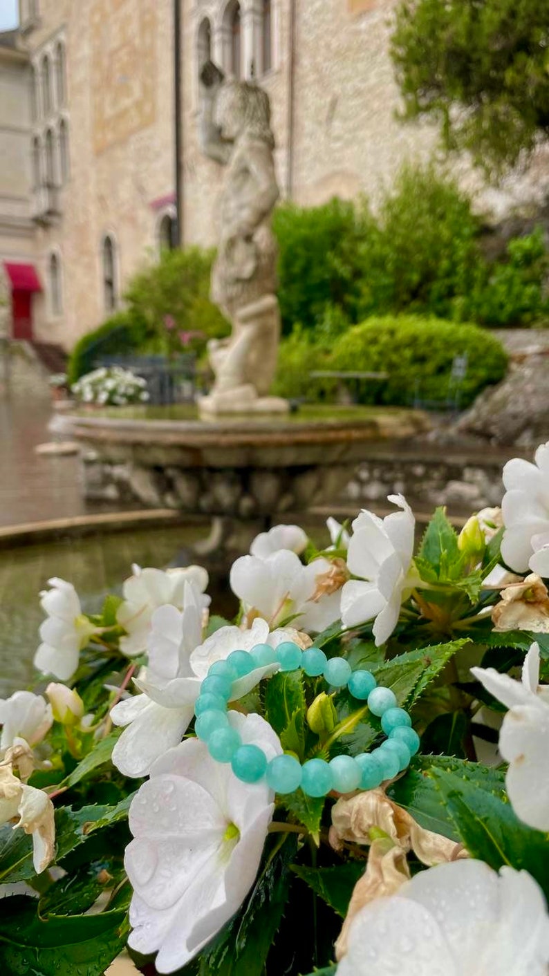 May include: A light blue beaded bracelet with round beads, resting on white flowers and green leaves. The bracelet is in focus, while the background is blurred and features a stone fountain and greenery.