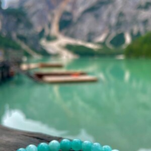 May include: A light blue gemstone bracelet with round beads. The bracelet is on a wooden surface with a blurry background of a lake and mountains.