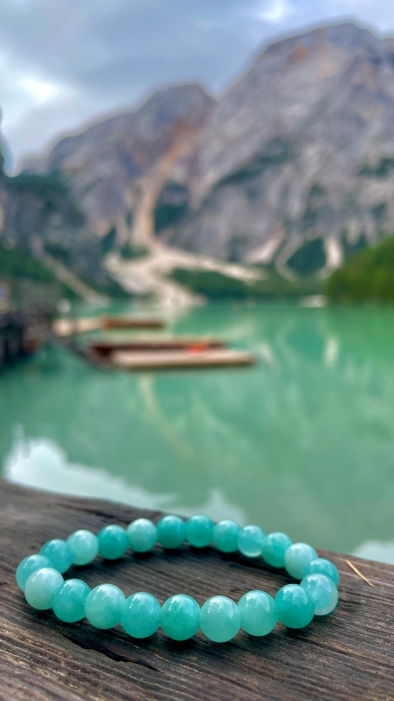 May include: A light blue gemstone bracelet with round beads. The bracelet is on a wooden surface with a blurry background of a lake and mountains.