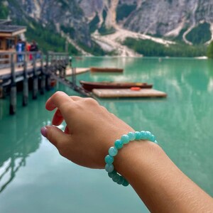 May include: A turquoise blue gemstone bracelet with round beads. The bracelet is worn on a person's wrist against a backdrop of a lake and mountains.
