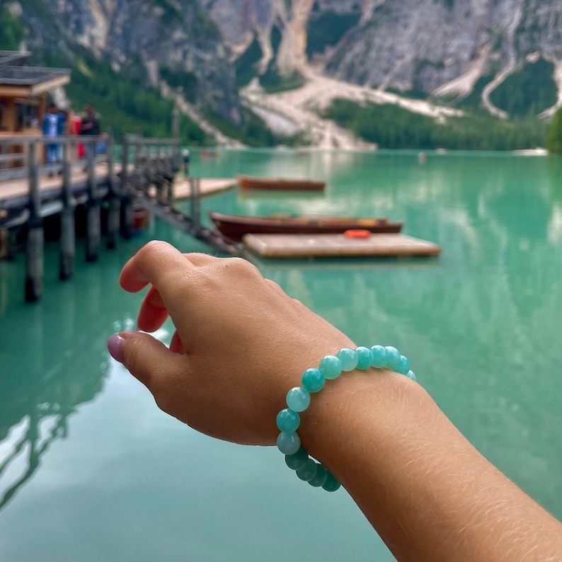 May include: A turquoise blue gemstone bracelet with round beads. The bracelet is worn on a person's wrist against a backdrop of a lake and mountains.