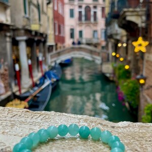 May include: A light blue gemstone bracelet with round beads. The bracelet is lying on a light brown textured surface. The background is a blurry image of a canal in Venice, Italy.