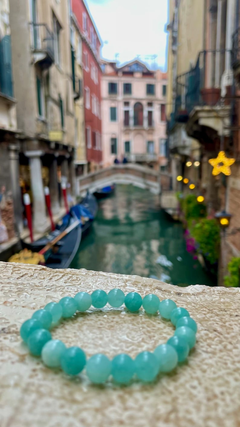 May include: A light blue gemstone bracelet with round beads. The bracelet is lying on a light brown textured surface. The background is a blurry image of a canal in Venice, Italy.