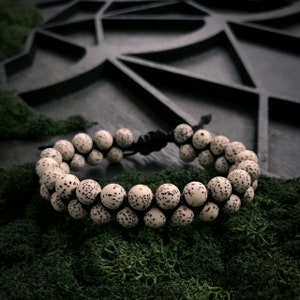 May include: A white beaded bracelet with black speckles, made of natural seeds, with a black cord closure. The bracelet is on a bed of green moss.