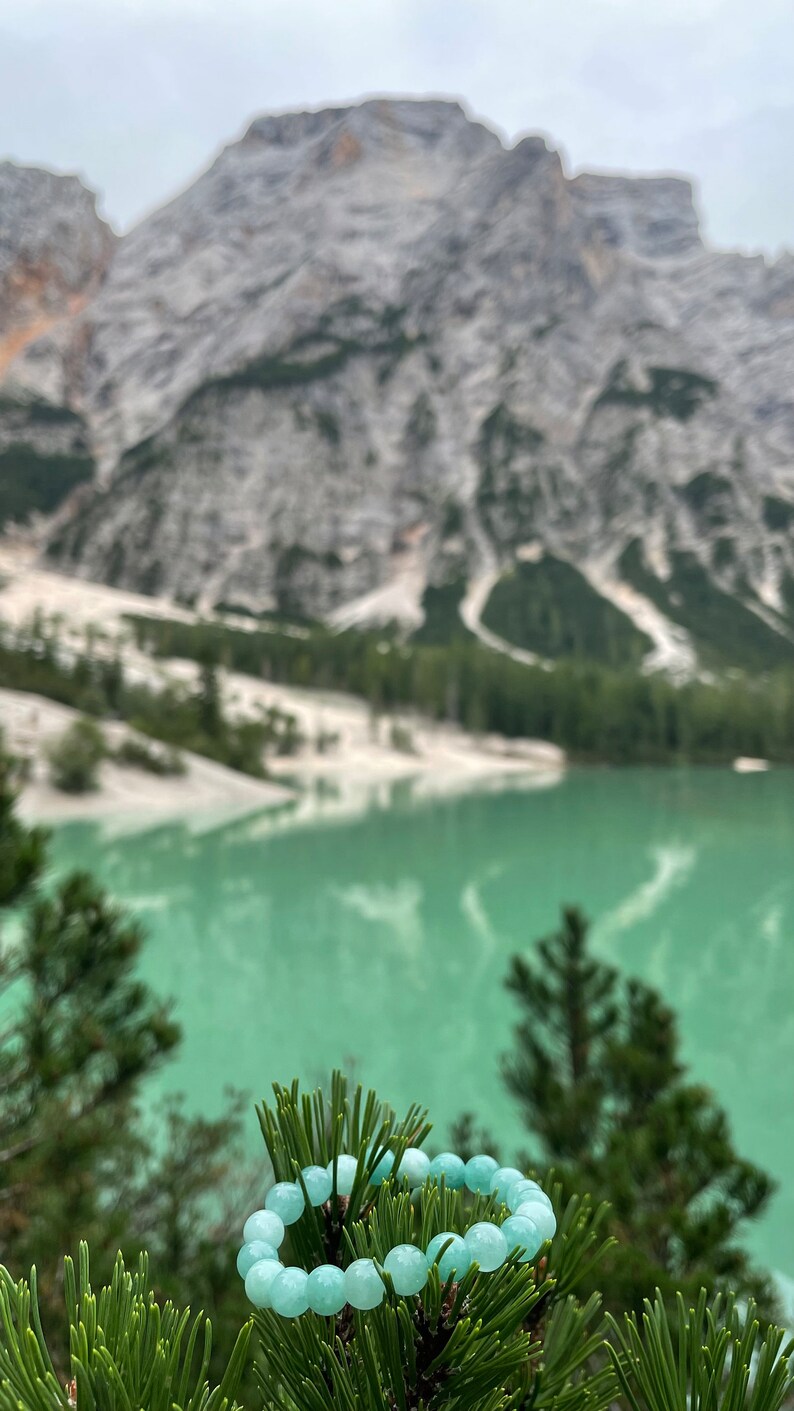 May include: A light blue gemstone bracelet on a pine tree branch with a mountain lake and mountains in the background.
