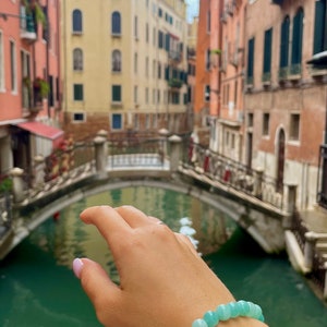 May include: A hand wearing a light blue gemstone bracelet with a canal and buildings in the background.