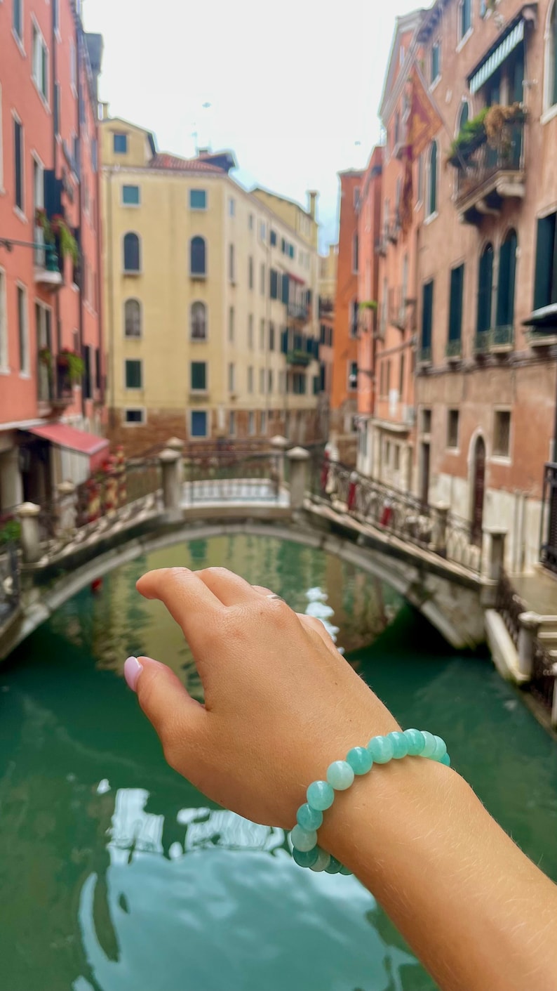 May include: A hand wearing a light blue gemstone bracelet with a canal and buildings in the background.