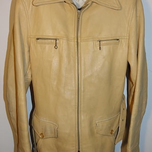 May include: A light tan leather jacket with a zip-up front, two chest pockets with small zipper pulls, and two lower pockets with button closures. The jacket has a wide collar and a belt detail on the sides.