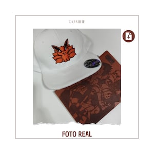 May include: A white baseball cap with a flat brim, featuring an orange and black embroidered fox design. A brown notebook with a detailed illustration is next to the cap. The text "FOTO REAL" is at the bottom.