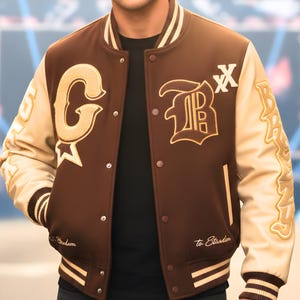 Könnte beinhalten: Braune und beige Collegejacke mit den Buchstaben "G" und "B" auf der Vorderseite gestickt. Die Jacke hat einen schwarzen Kragen und Bündchen mit weißen Streifen. Die Ärmel sind beige und der Körper ist braun. Auf der Jacke stehen die Worte "Bredum" und "To Stardom".